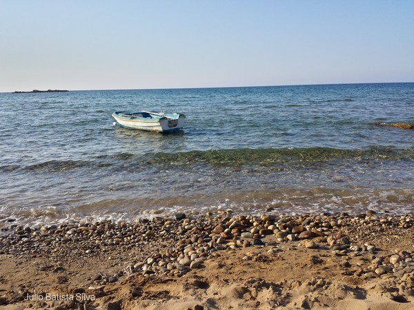 A small white boat floats in calm blue water near a pebble-strewn shore under a clear sky.