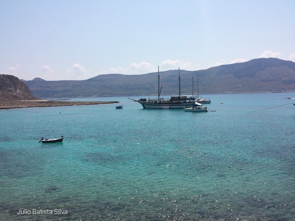 A large sailboat and smaller boats are anchored in the clear turquoise water of a coastal bay, with mountains in the background.