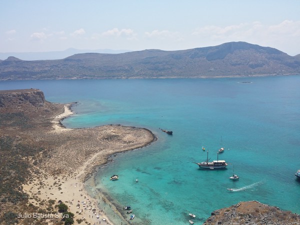 An aerial photograph captures a scenic coastal bay with turquoise water, a sandy beach, and several boats near a rugged shoreline.