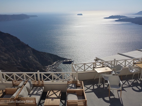A scenic view from a terrace overlooking the Aegean Sea, with white furniture and a distant island on the horizon.