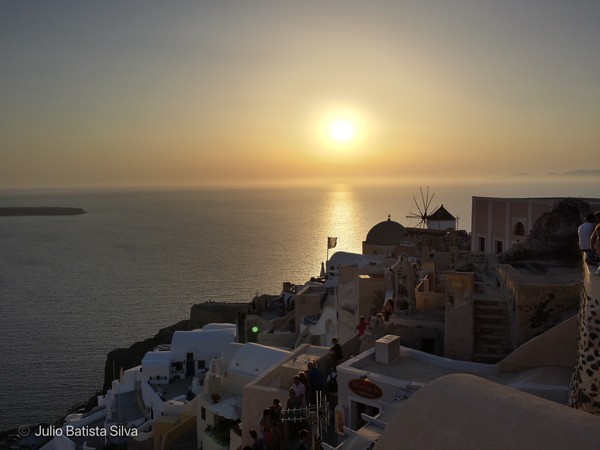 A stunning sunset over the Aegean Sea, casting a golden glow on the white-washed buildings and a traditional windmill of a Greek island village.