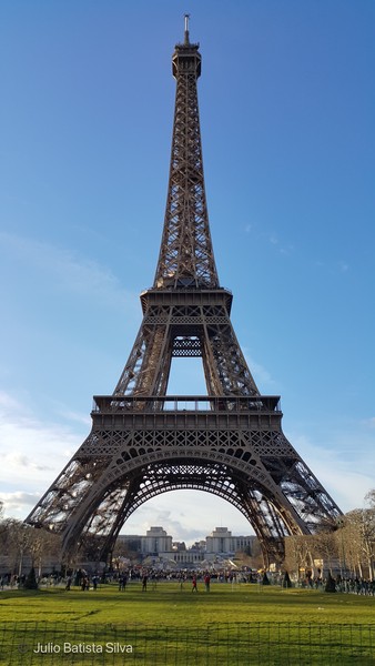 A low-angle view captures the Eiffel Tower against a clear blue sky, with people on the grassy foreground.