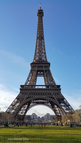 A low-angle view captures the Eiffel Tower against a clear blue sky, with people on the grassy foreground.