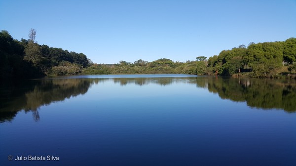 A serene lake reflects the surrounding trees and a clear blue sky, creating a peaceful natural landscape.