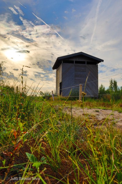 A low-angle shot captures a wooden cabin in a grassy field under a dramatic sky with clouds and contrails.