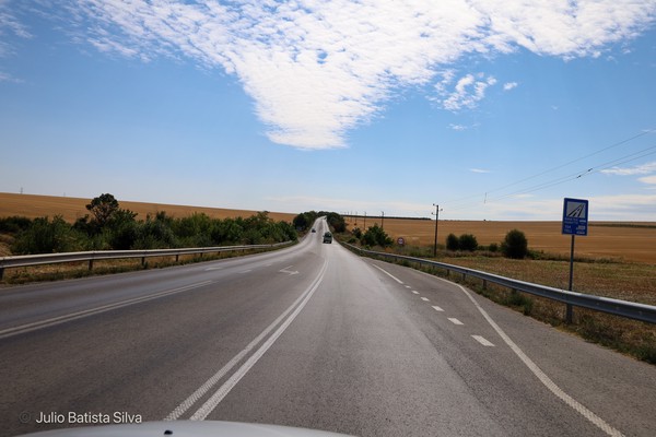 A long, winding road cuts through a rural landscape of golden fields under a bright blue sky with scattered clouds.