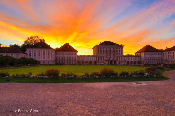 A grand palace is silhouetted against a vibrant sunset sky, with a large green lawn in the foreground.