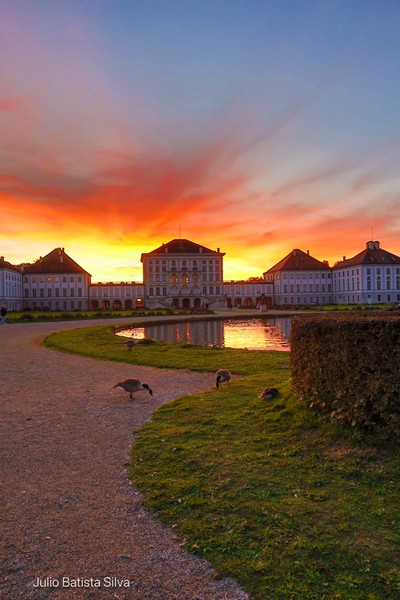 A grand palace is silhouetted against a vibrant sunset, with geese grazing on the grassy grounds.