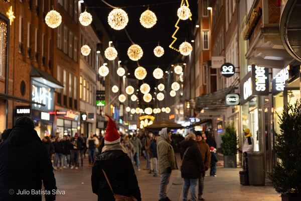 A crowded city street at night, illuminated by hanging spherical lights and a person wearing a red Santa hat.