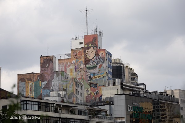 A large, colorful graffiti mural of a girl adorns the side of a building in a city under a cloudy sky.