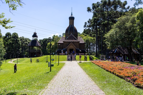 A wooden church with a dome stands on a grassy hill, surrounded by trees and a colorful flower bed, with people walking on a path.