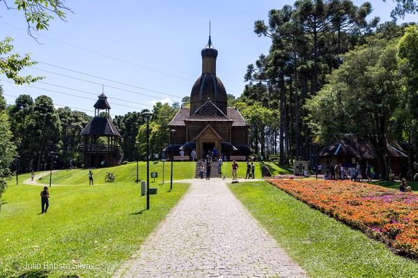 A wooden church with a dome stands on a grassy hill, surrounded by trees and a colorful flower bed, with people walking on a path.