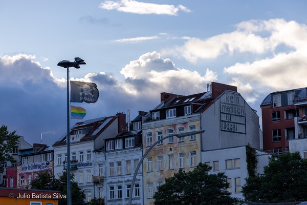 A cityscape with a large mural reading 'kein mensch ist illegal' and a flag with a skull and crossbones.