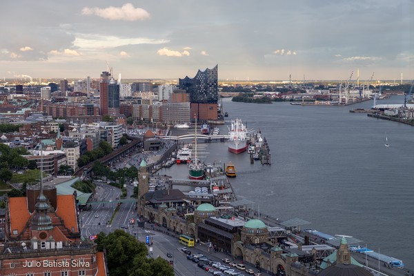 An aerial photograph captures the Hamburg harbor at dusk, featuring the Elbphilharmonie, a large ship, and surrounding city architecture.