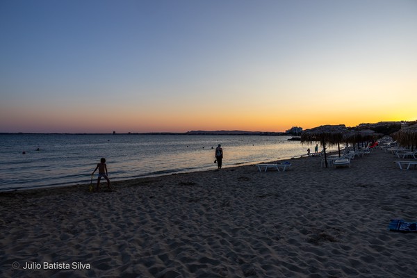 A serene beach scene at dusk, with silhouetted figures playing in the sand and water against a vibrant orange and blue sky.