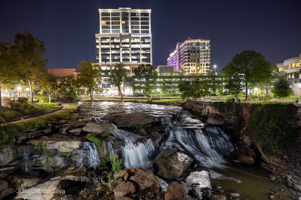A long-exposure photograph captures a cascading waterfall in an urban park at night, with illuminated buildings in the background.