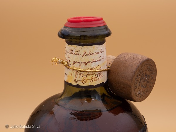 A close-up of a vintage dark glass bottle with a red wax seal, a handwritten paper label, and a wooden stopper.
