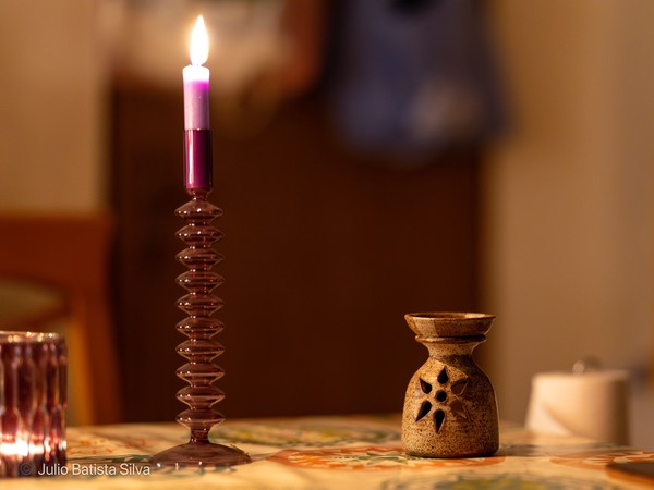 A lit purple candle in a glass holder casts a warm glow on a patterned table, with a ceramic oil burner nearby.
