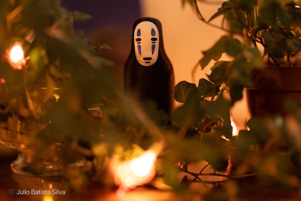 A No-Face figurine is centered in a dimly lit scene, surrounded by green foliage and warm, glowing lights.
