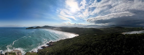 A panoramic view of a coastline with a forested hillside, ocean waves, and a dramatic sky.